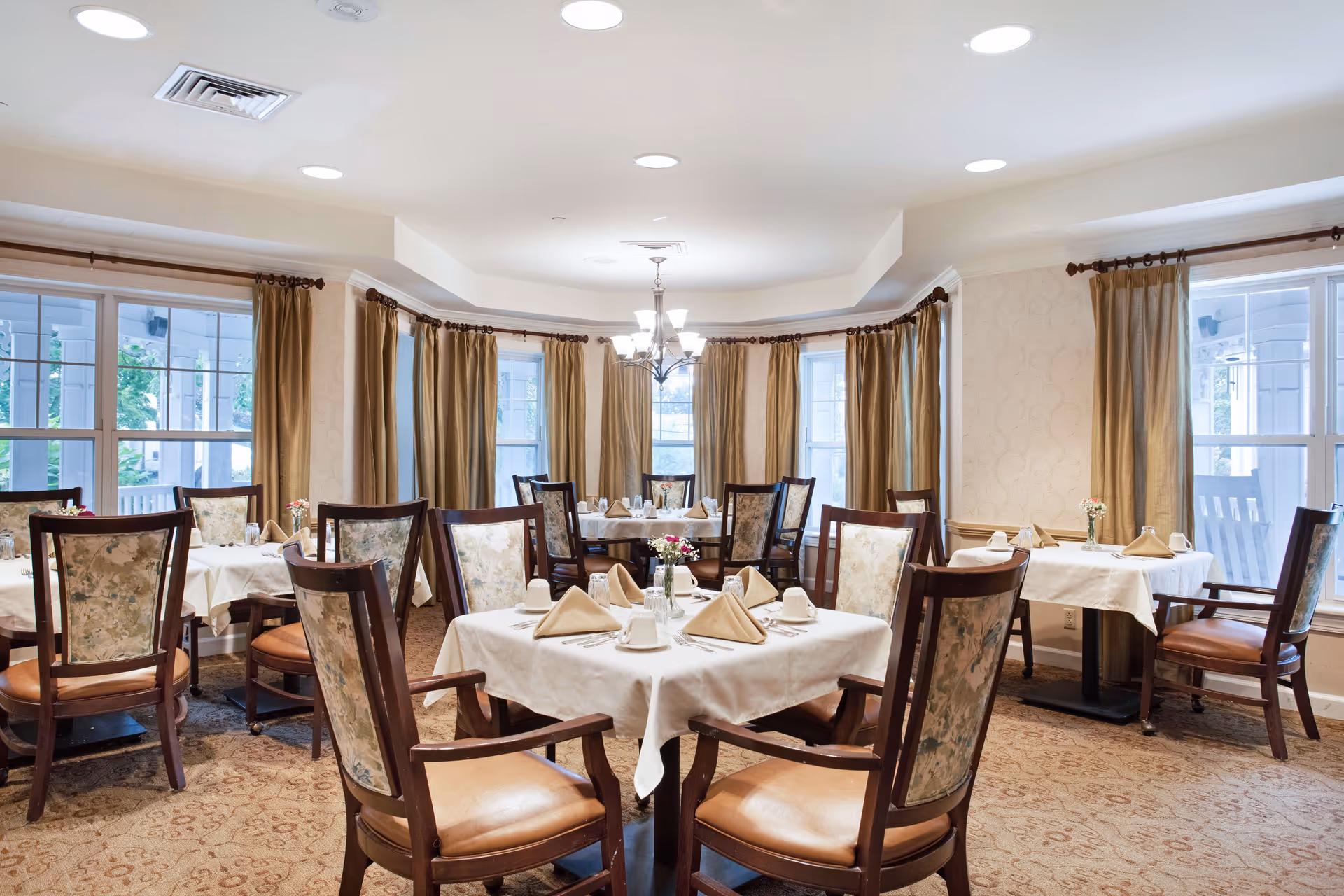 A bright dining room with multiple tables covered in white tablecloths, each set with beige napkins, cups, and silverware. The room features large windows with gold curtains, floral upholstered wooden chairs, a patterned carpet, and a chandelier hanging from the ceiling.