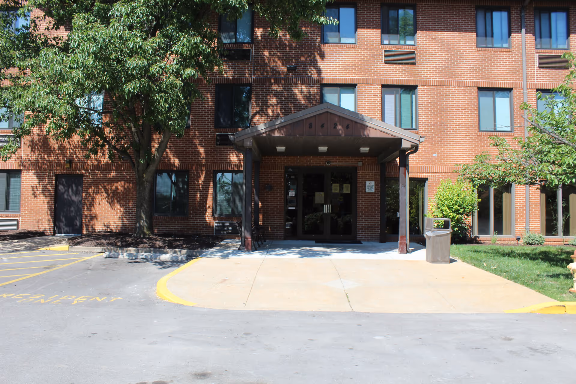 Front entrance of a red-brick apartment building with a covered entryway, trees, windows, and a small driveway.