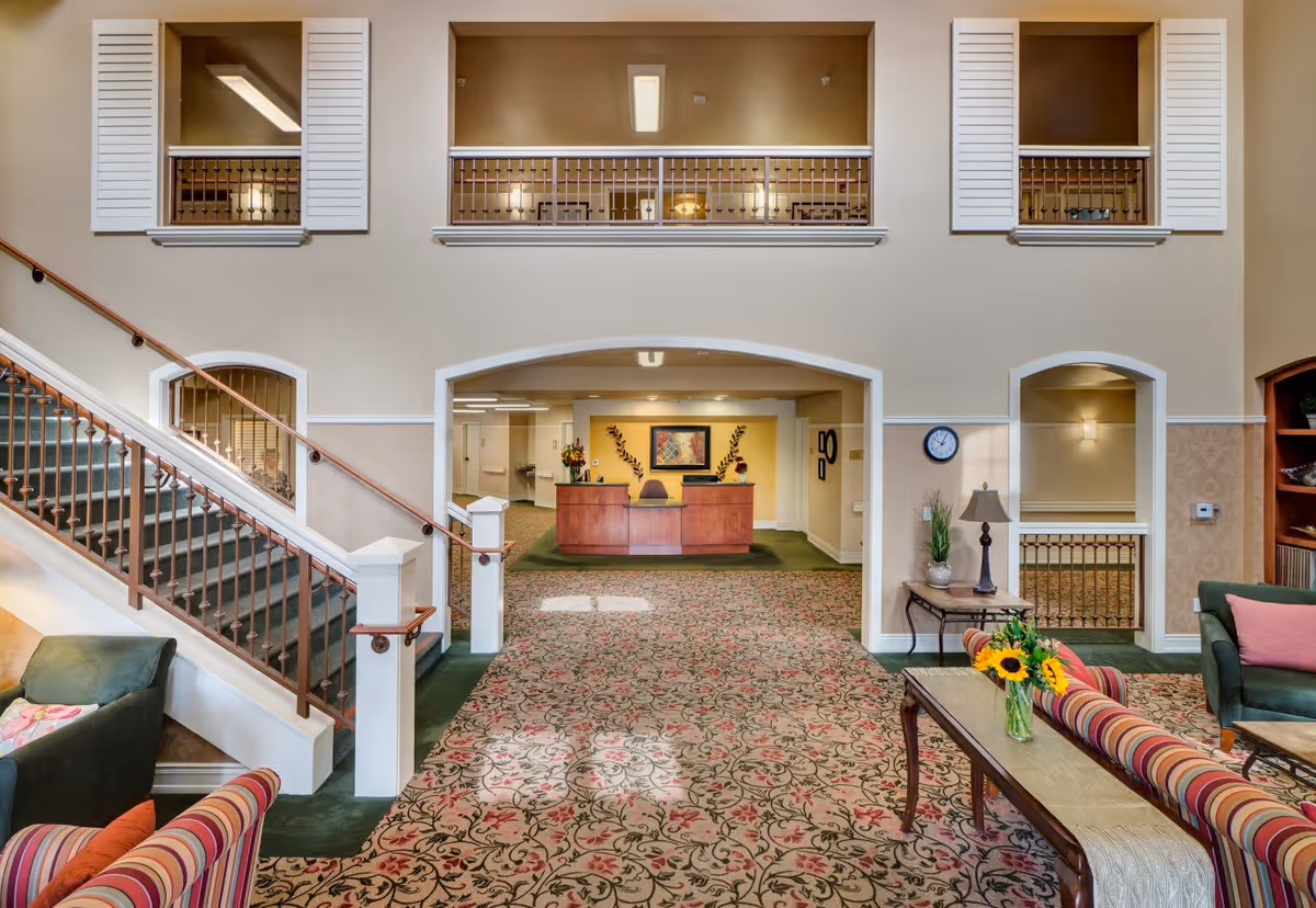Interior view of a senior living facility lobby area with a patterned carpet, a wooden reception desk against a yellow accent wall, and a staircase with a brown railing on the left. The room has high ceilings with windows and railings on the upper level. There are comfortable seating areas with striped and solid-colored sofas and a coffee table with a vase of sunflowers.