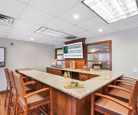 U-shaped granite countertop bar with wooden high-back chairs and a sign reading "Bentley Commons at Paragon Village" on the back wall.