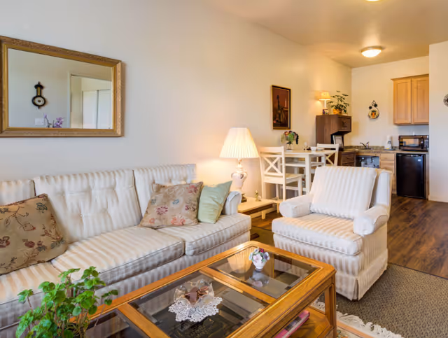 A cozy living room area with a striped white sofa and matching armchair, decorated with floral and solid pillows. A wooden coffee table with glass panels holds a small flower vase and decorative items. A table lamp on a side table provides warm lighting. In the background, there is a small dining table with two chairs, a wooden cabinet, and a kitchenette with a sink, microwave, and mini fridge. The room has wooden flooring and light-colored walls with framed artwork and a mirror.