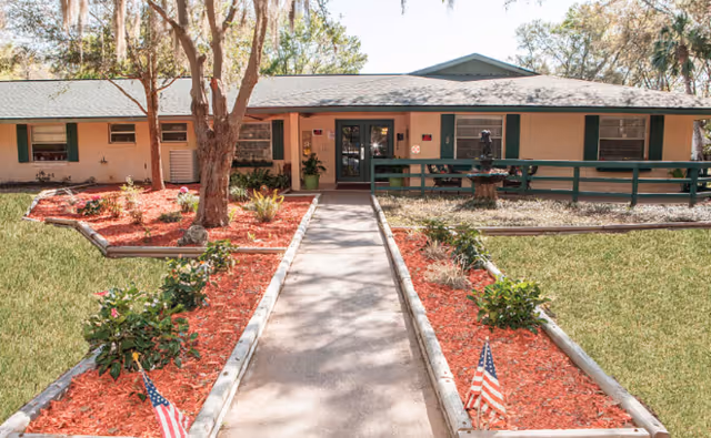 Single-story beige building with green trim and a central walkway through landscaped mulched beds leading to the main entrance.