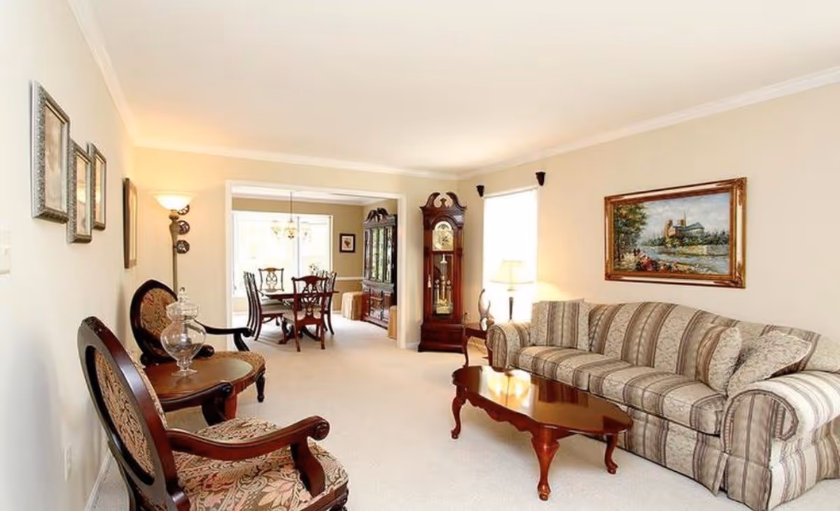 Bright traditional living room with a striped sofa, wooden coffee table, armchairs, a grandfather clock, and a view into a dining area.