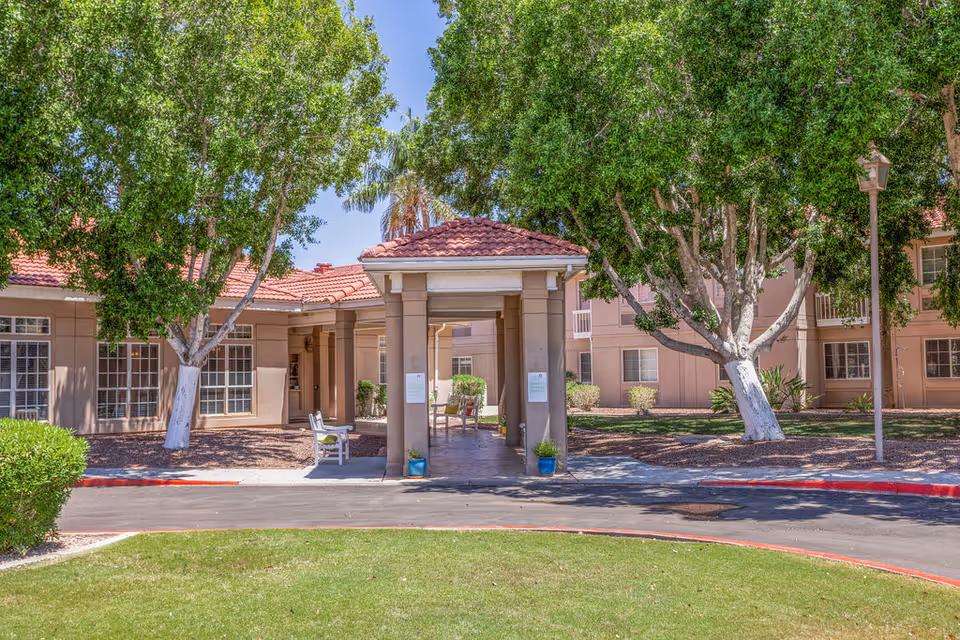 The entrance of The Gardens of Sun City, featuring a covered porch with seating, surrounded by lush greenery and a well-maintained landscape.