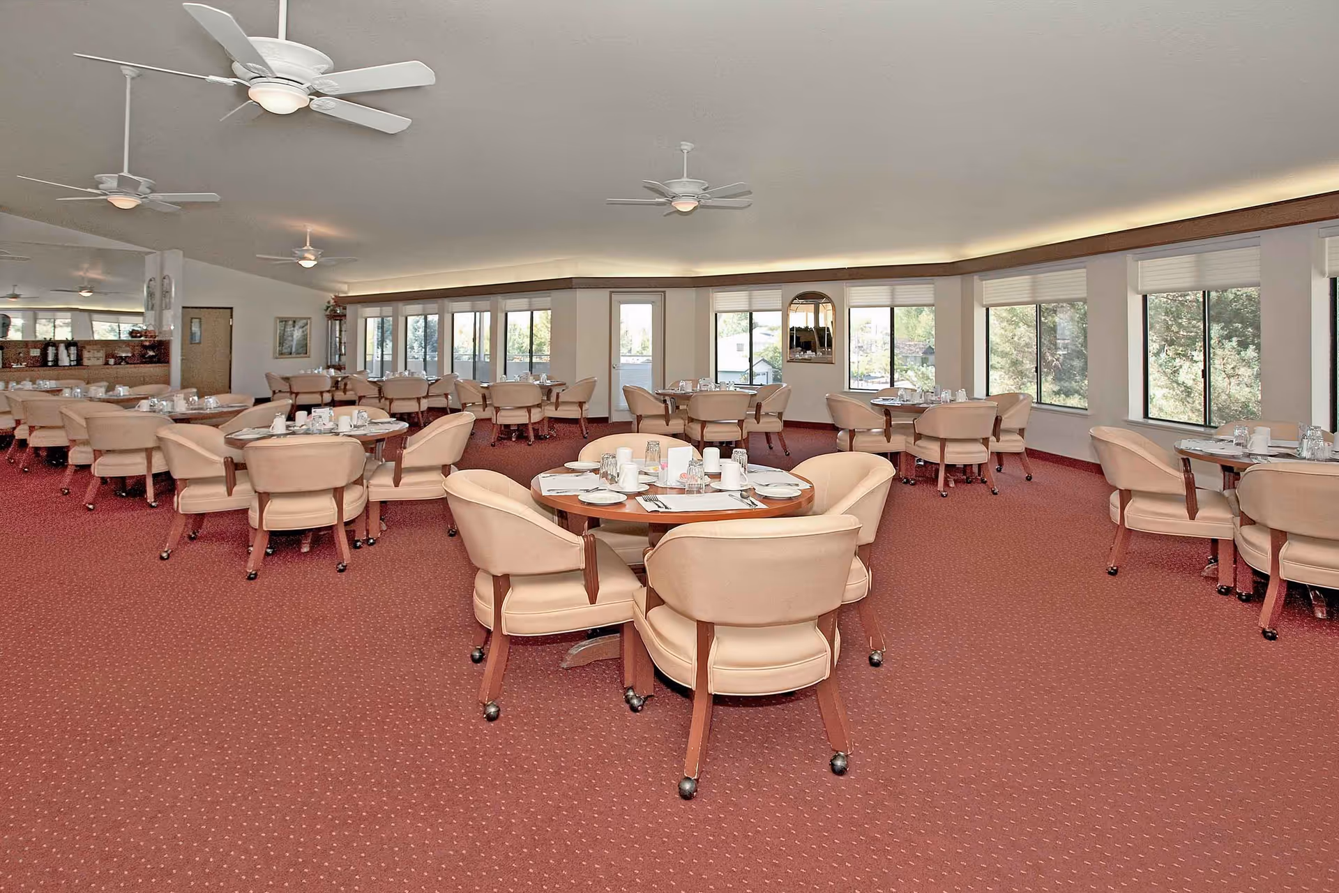 A spacious dining room with multiple round tables set with white plates, cups, and silverware. The room has beige cushioned chairs with wooden legs on casters, red carpet flooring, large windows letting in natural light, ceiling fans, and a mirrored wall on one side.