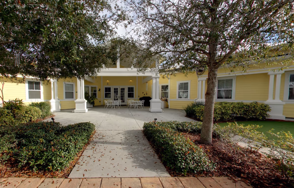 Front entrance of a yellow senior living building with a covered patio, white columns, outdoor tables, and landscaped shrubs and trees.