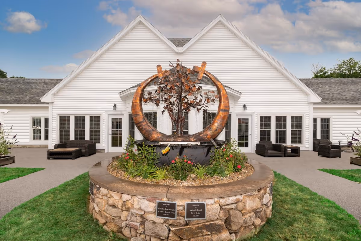 Outdoor view of a white building with multiple windows and doors, featuring a circular stone planter with a metal sculpture of a tree and large curved elements in the center. There are patio chairs and tables on either side of the building entrance, with green grass and a partly cloudy sky in the background.