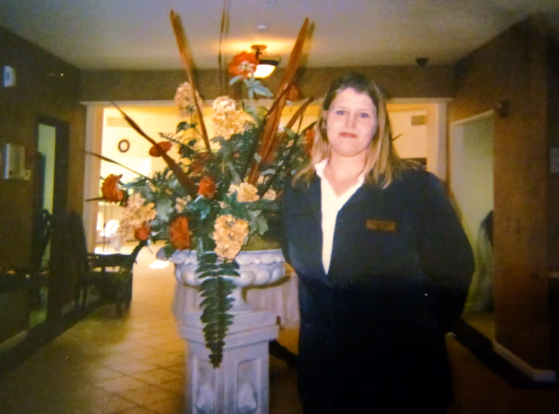 A woman with light brown hair wearing a dark blazer and white shirt stands indoors next to a large decorative floral arrangement in a white ornate vase. The background shows a hallway with warm lighting and some furniture.