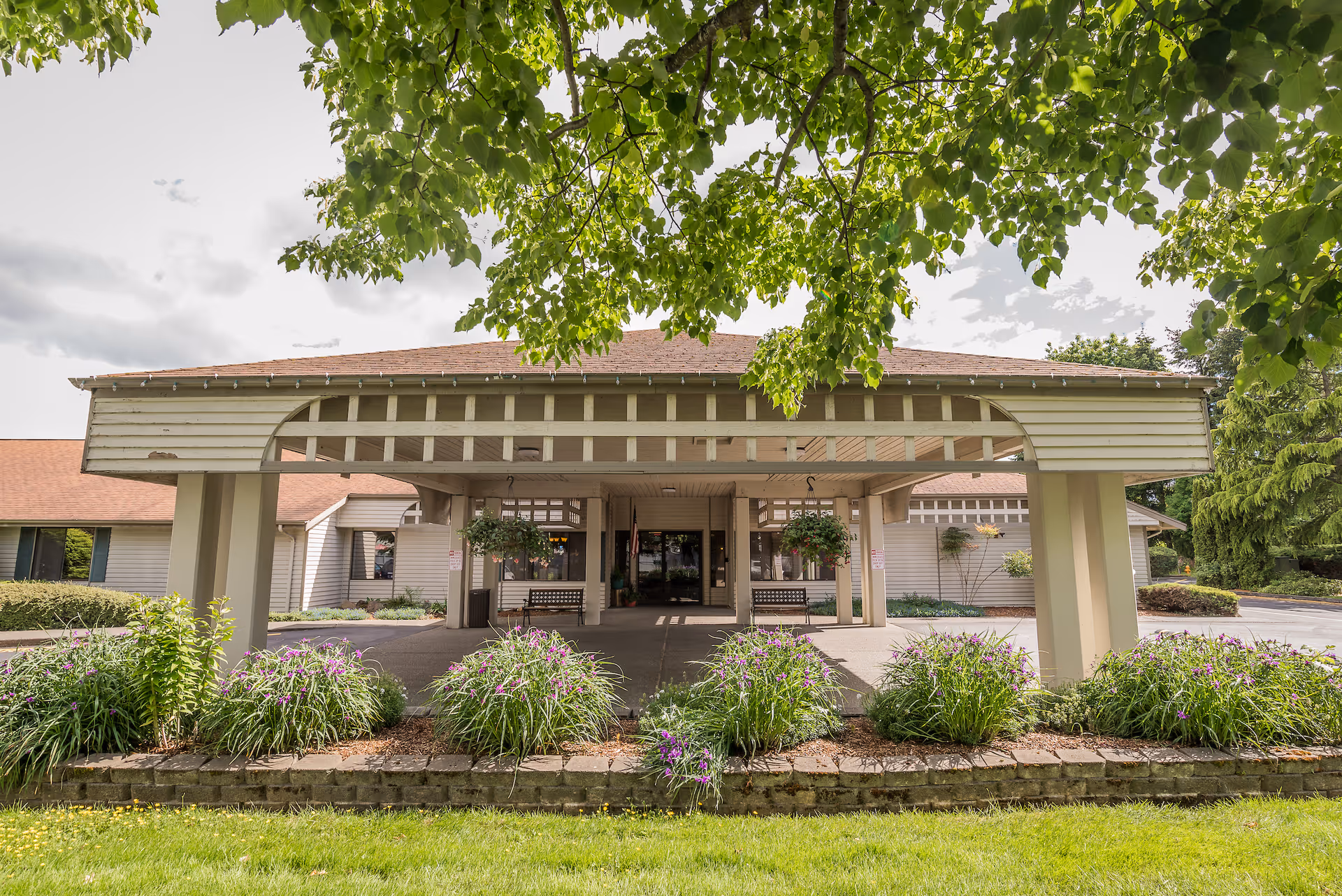 Front entrance of Village Concepts of Marysville - Grandview Village with a covered driveway, hanging flower baskets, benches, and landscaped greenery including bushes and trees.