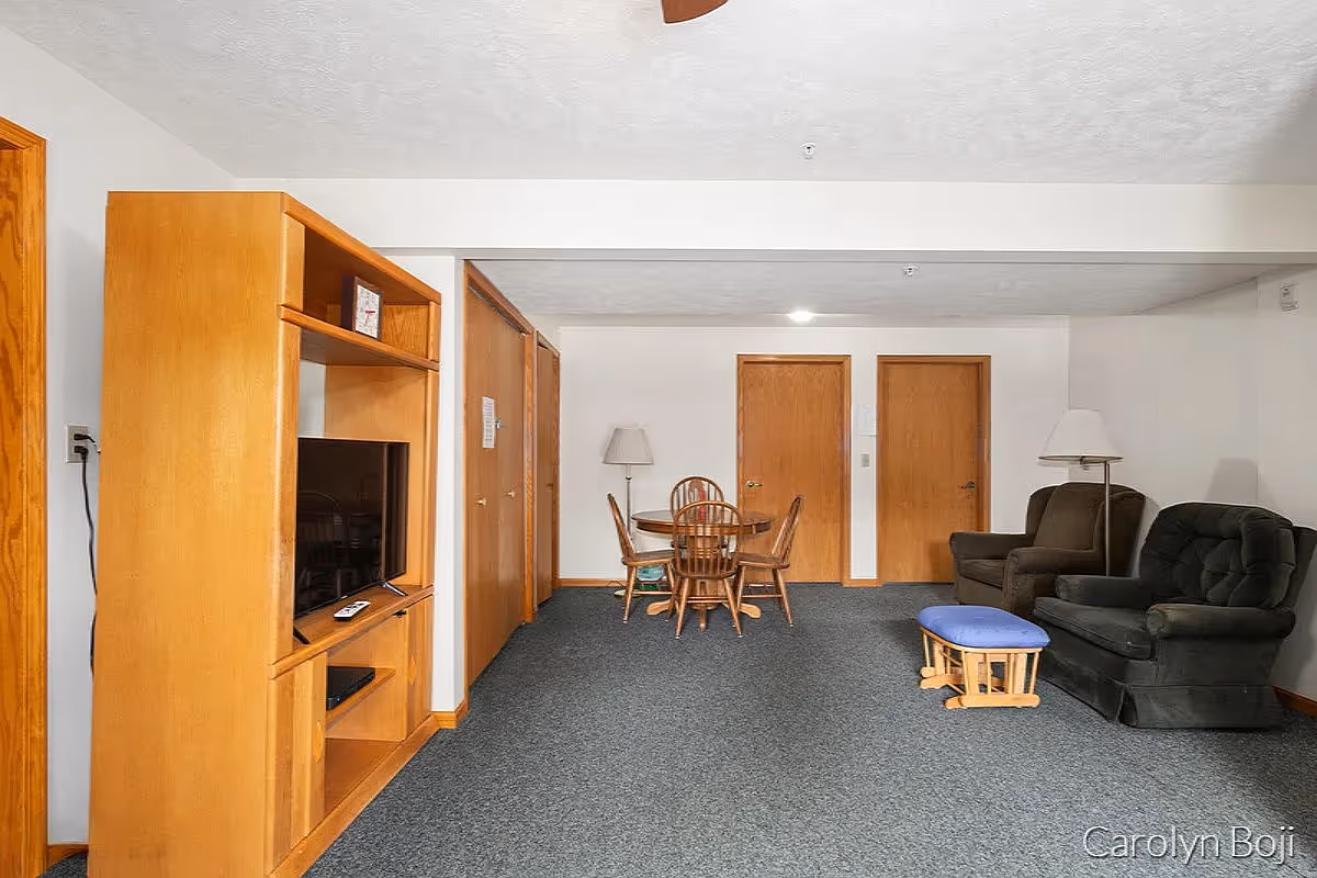 A living room area with a wooden entertainment center holding a TV on the left, a round wooden dining table with four chairs in the center, and two upholstered armchairs with a blue cushioned footstool on the right. The room has gray carpet, white walls, and wooden doors in the background. Two floor lamps are positioned near the chairs.