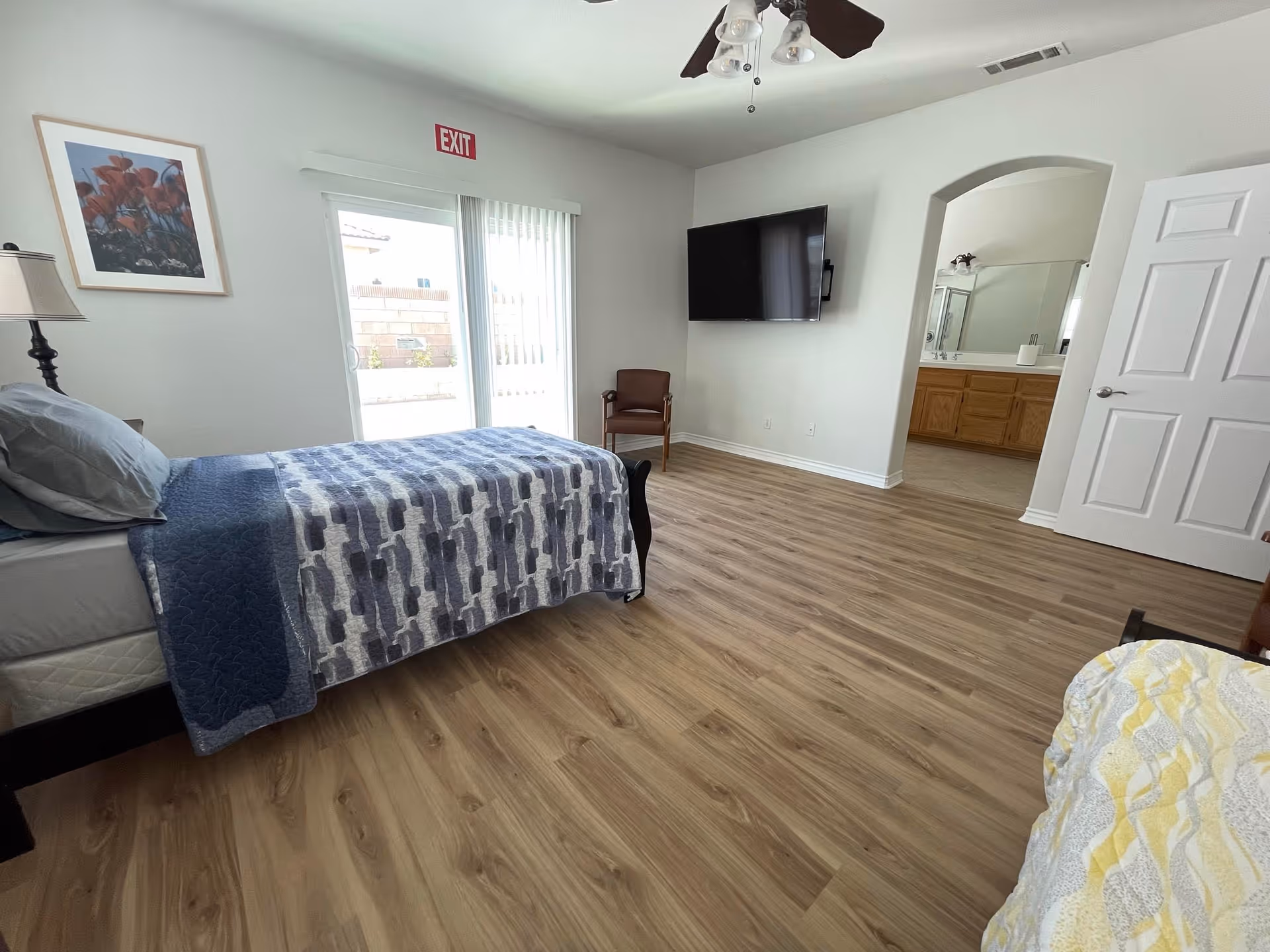 A bright bedroom with a single bed covered in a blue and gray patterned quilt, a wooden floor, a wall-mounted flat-screen TV, a brown chair, a ceiling fan with lights, a framed flower picture on the wall, and a sliding glass door with vertical blinds leading outside. An open doorway reveals a bathroom with a large mirror and wooden cabinets.