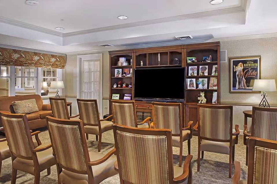 Comfortable common room with rows of chairs facing a large television set in a wooden entertainment unit and seating area.