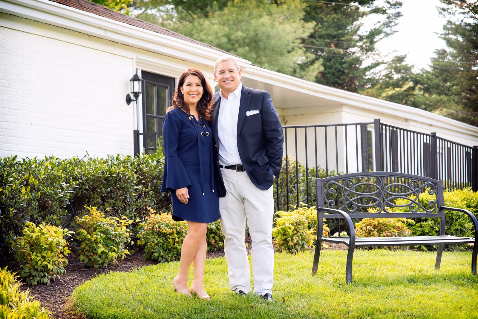 A man and woman standing together outside near a white brick building with black railings and green bushes, smiling at the camera. There is a black metal bench on the grass nearby.