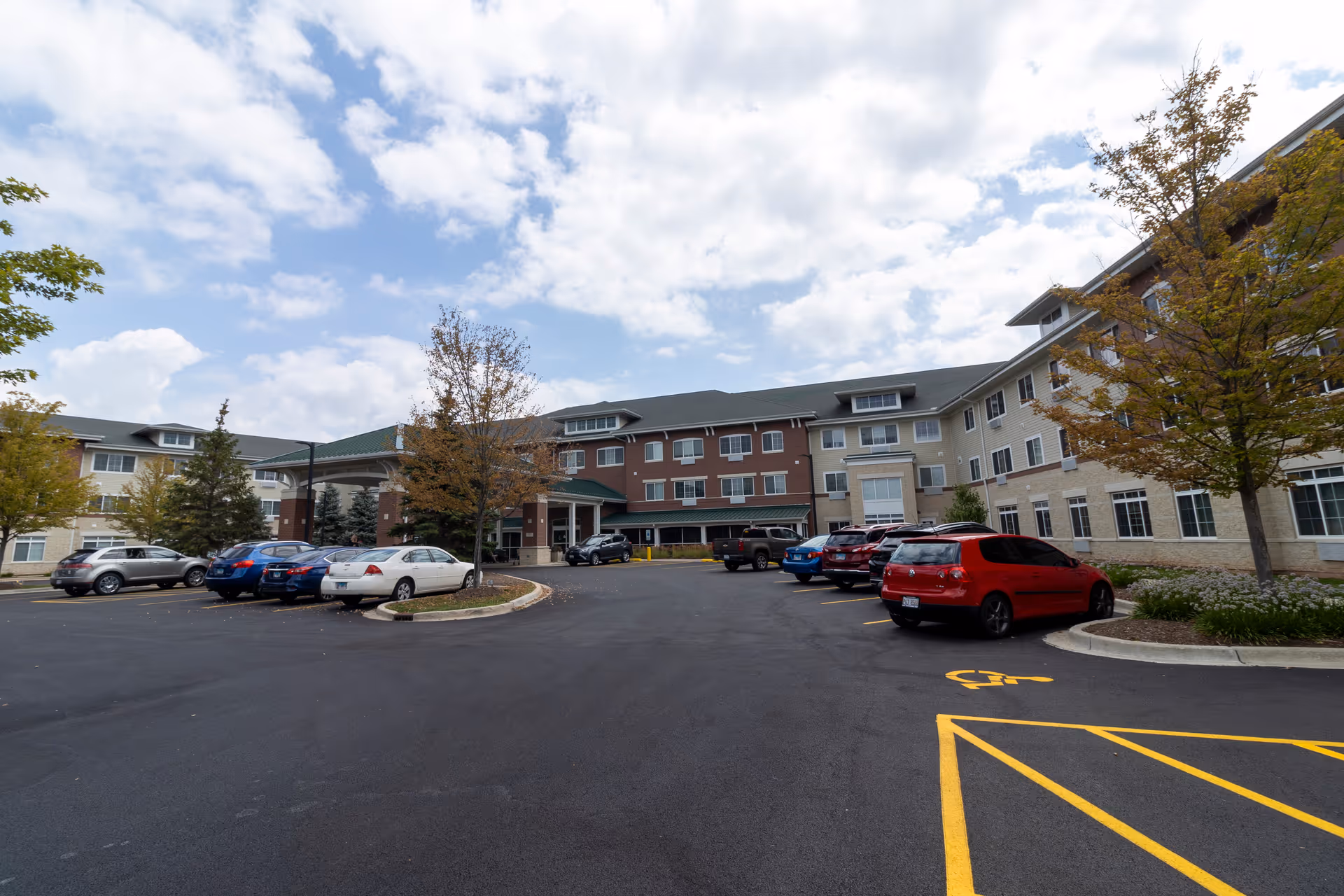 Exterior view of a multi-story senior living facility building with a parking lot in front. Several cars are parked, and there are trees with autumn foliage around the lot. The sky is partly cloudy.