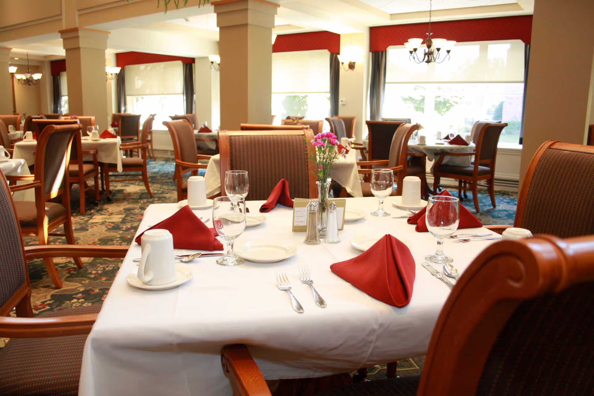 A dining room in a senior living facility with tables set for a meal. Each table is covered with a white tablecloth and has red folded napkins, glassware, plates, and silverware. There are wooden chairs with cushioned seats around the tables. The room has large windows letting in natural light and decorative light fixtures on the walls and ceiling.