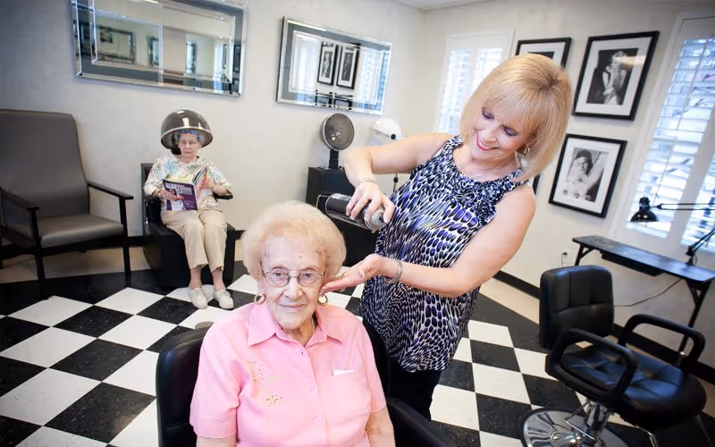 A hair salon inside a senior living facility where a woman is styling the hair of an elderly woman seated in a salon chair. Another elderly woman is sitting under a hair dryer in the background, reading a magazine. The room has black and white checkered flooring, mirrors on the wall, and framed black and white photos.