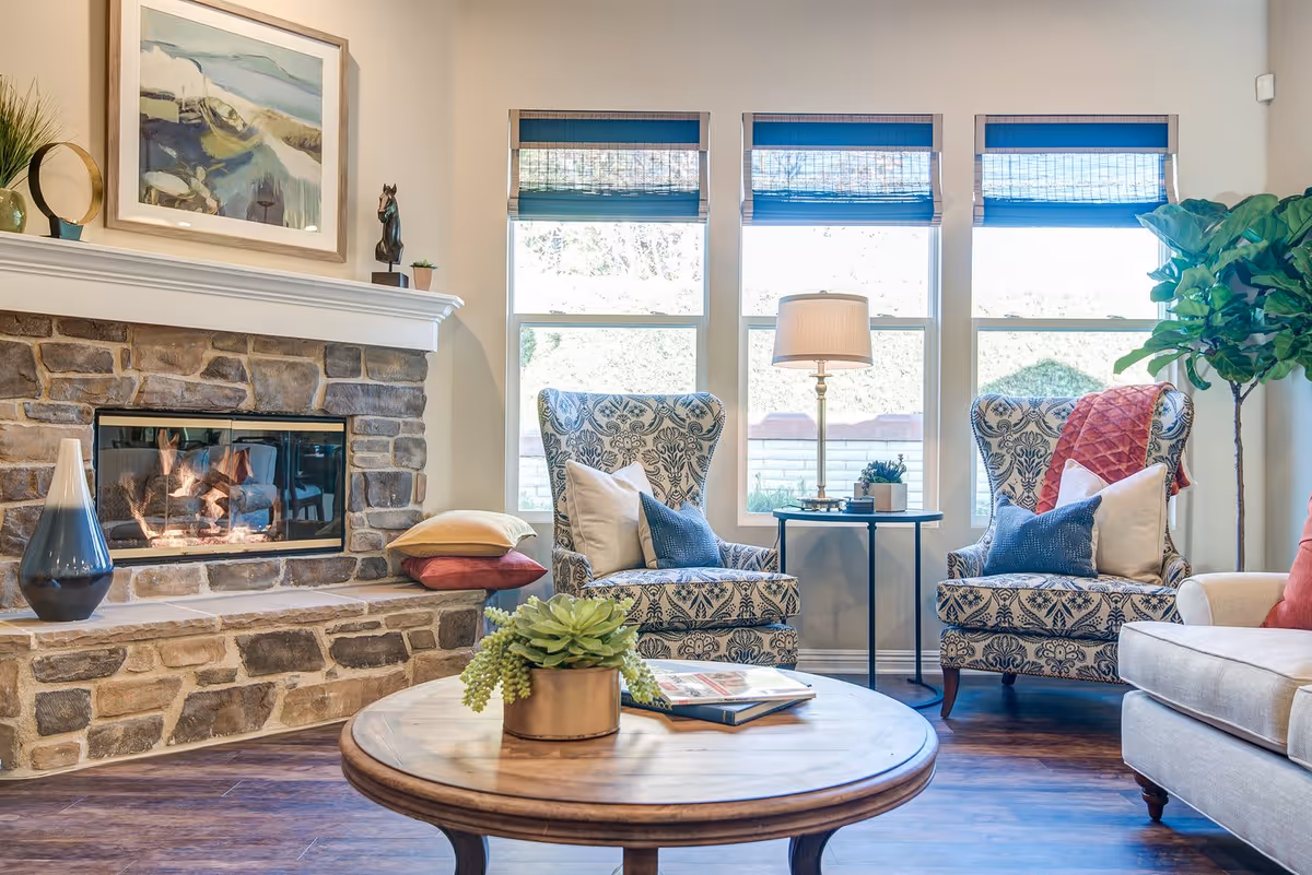 A cozy living room with a stone fireplace on the left, a round wooden coffee table with a small plant and books, two patterned armchairs with pillows and a side table with a lamp between them, and a beige sofa on the right. Three large windows with blue blinds let in natural light, and there is a tall green plant in the corner.