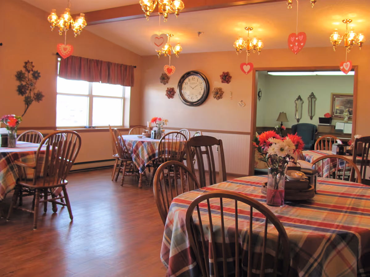 Dining room with plaid-covered tables, wooden chairs, chandeliers, and floral centerpieces.