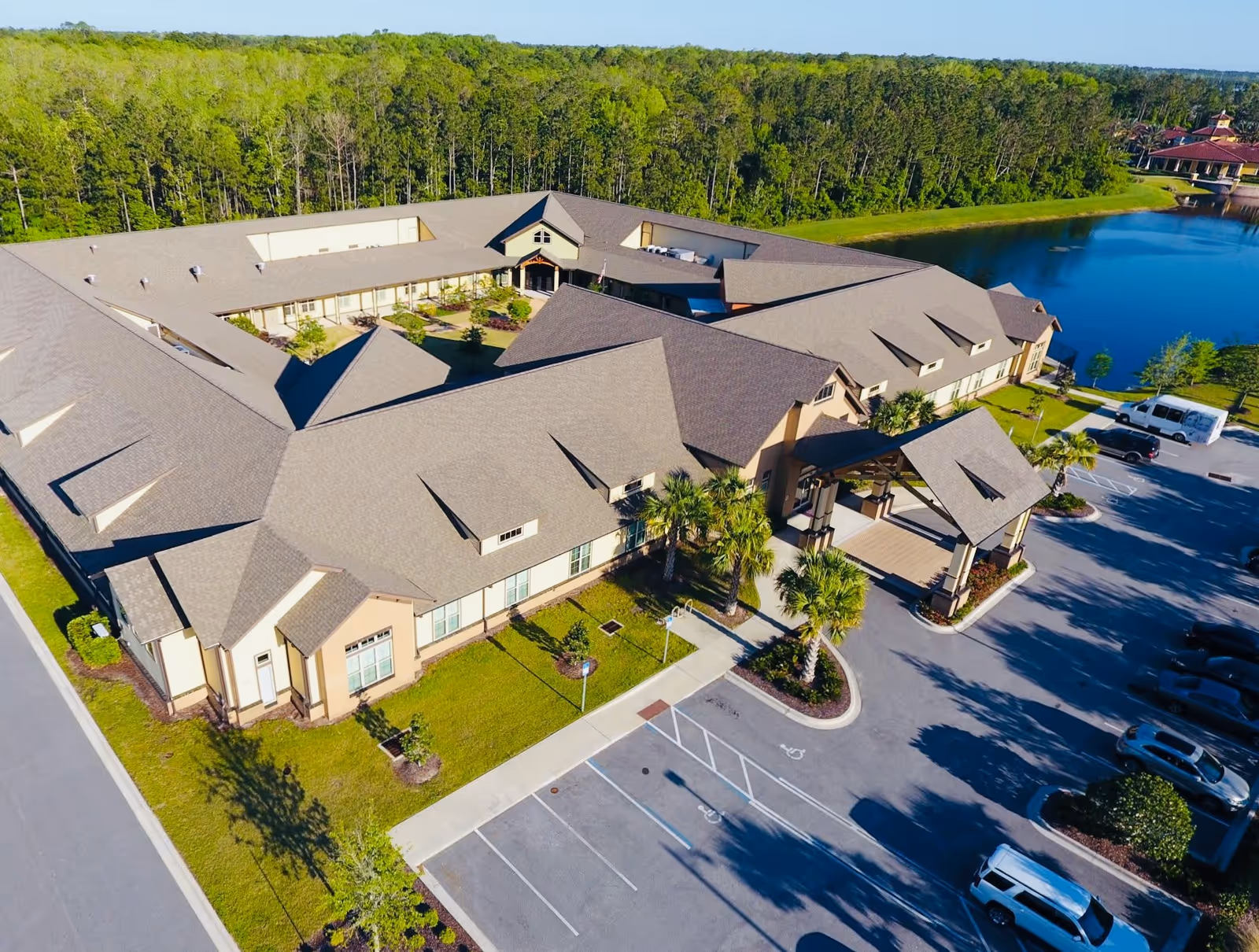 Aerial view of Rosecastle at Deerwood senior living facility showing a large building with a courtyard, surrounded by trees and a pond. The parking lot in front has several cars parked and a covered entrance with palm trees nearby.