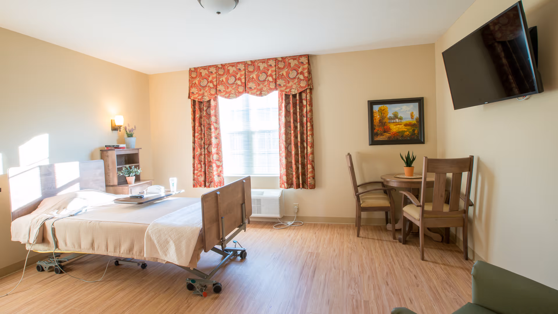 A bright and clean nursing center bedroom with a hospital-style bed, a small wooden table with two chairs, a wall-mounted TV, a window with patterned red curtains, and light wood flooring.