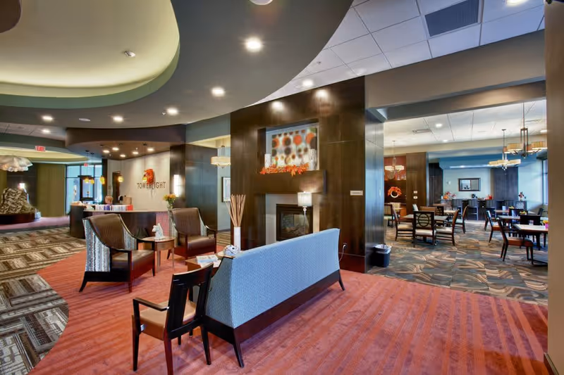 Interior view of a senior living facility lobby and dining area with modern furniture, a fireplace, and decorative elements including a floral arrangement and artwork. The space features a mix of carpet patterns and ceiling lights, with a reception desk visible in the background.