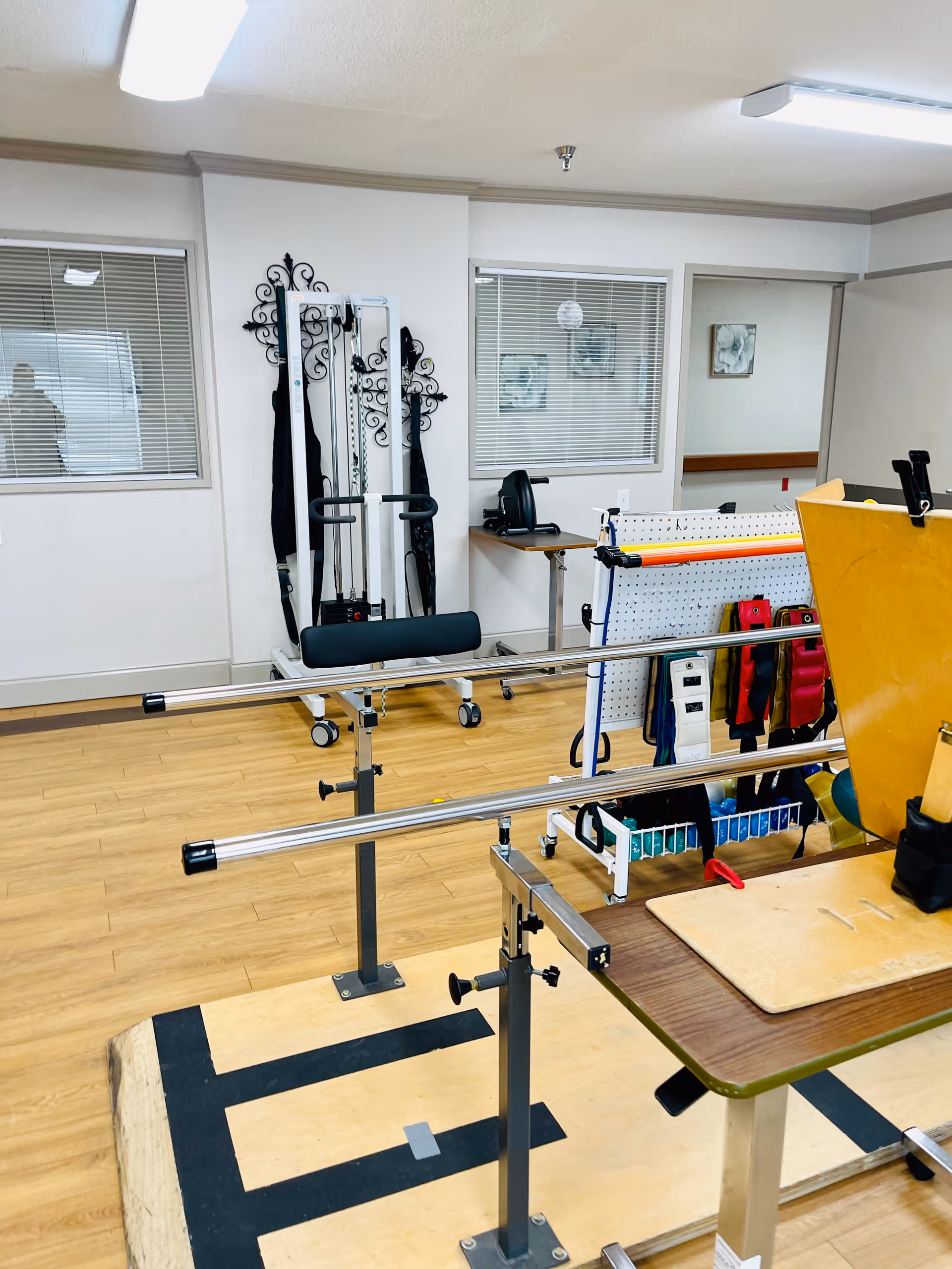 Physical therapy room with parallel bars for walking exercises, exercise equipment, and a table with therapy tools. The room has wooden flooring, white walls, and windows with blinds.