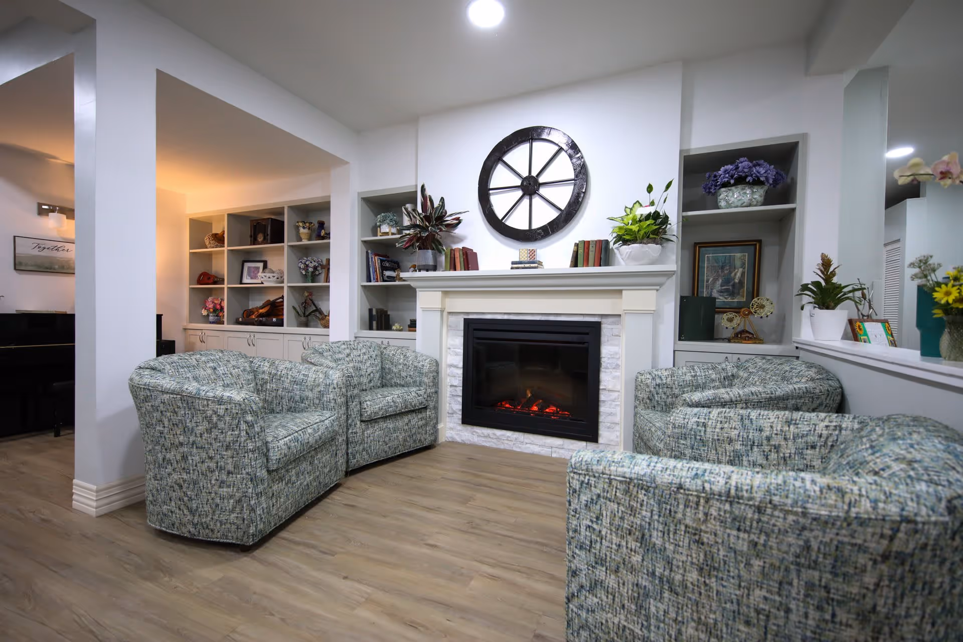 A cozy living room area with four patterned armchairs arranged around a white fireplace with a black electric insert. Above the fireplace is a large decorative clock. Built-in shelves on either side of the fireplace hold plants, books, and decorative items. The room has light wood flooring and white walls, with a piano visible in an adjacent space.
