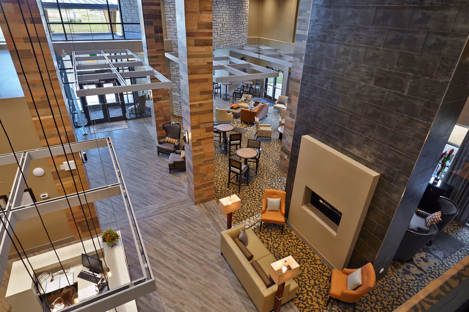 View from above of a spacious senior living facility common area with a modern fireplace, multiple seating arrangements including sofas and armchairs, small round tables with chairs, and a reception desk with a person working. The area features wood and stone accents, large windows allowing natural light, and a patterned carpet.