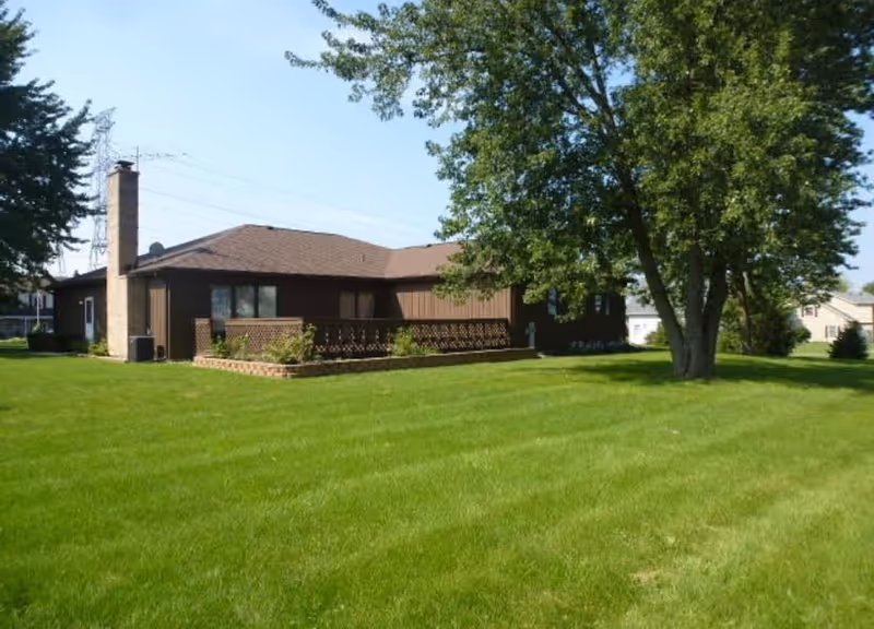 Single-story brown building with a chimney, surrounded by a well-maintained green lawn and several trees under a clear blue sky.