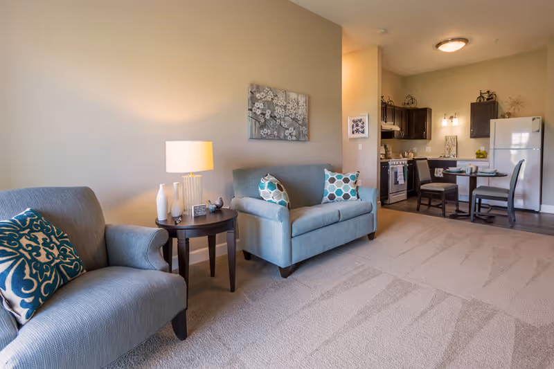 Open living area with two upholstered chairs and a loveseat around a side table, and an adjacent kitchen and dining area.
