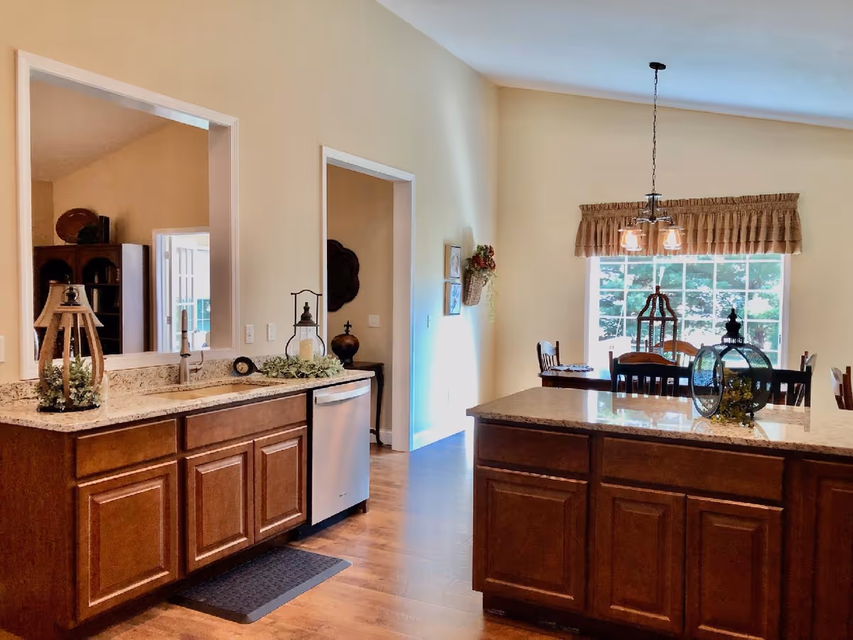 A bright kitchen and dining area with wooden cabinets and granite countertops. The kitchen features a sink, dishwasher, and decorative items on the counters. In the background, there is a dining table with chairs near a large window with a valance, allowing natural light to fill the room.