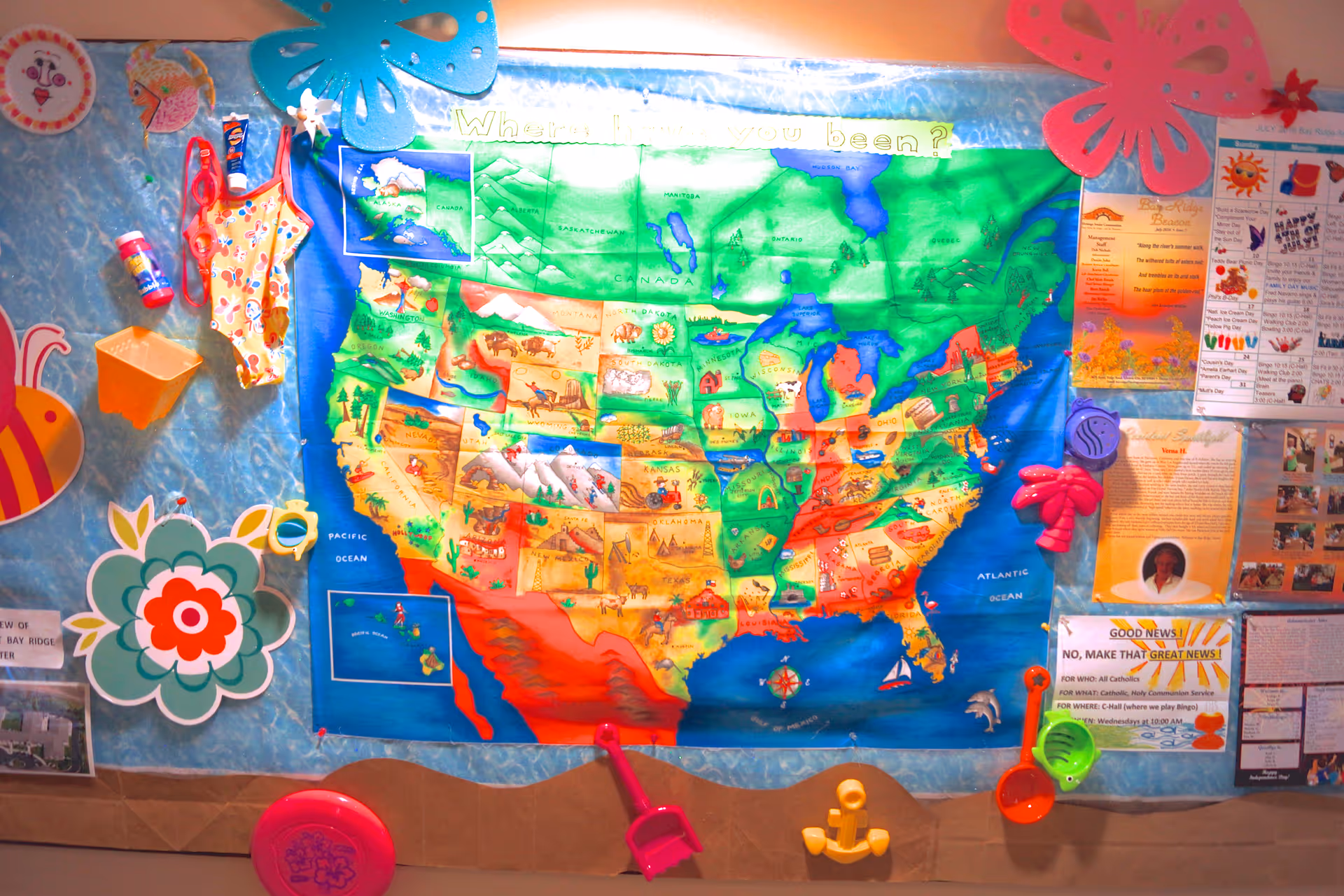 A colorful bulletin board featuring a large illustrated map of the United States with various decorations including paper flowers, a swimsuit, a small shovel, and other playful items. There are also several notices and flyers pinned around the map, one of which mentions 'Good News' and community activities.