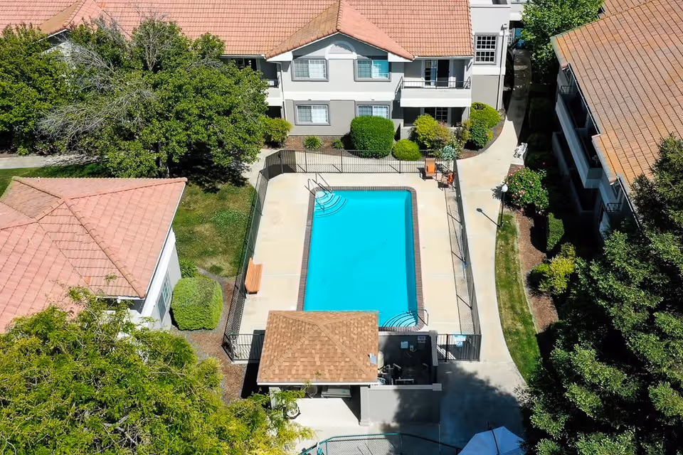 Aerial view of a rectangular swimming pool surrounded by a concrete deck and black metal fence, located in the courtyard of a senior living facility with multiple two-story buildings with red-tiled roofs and green landscaping.