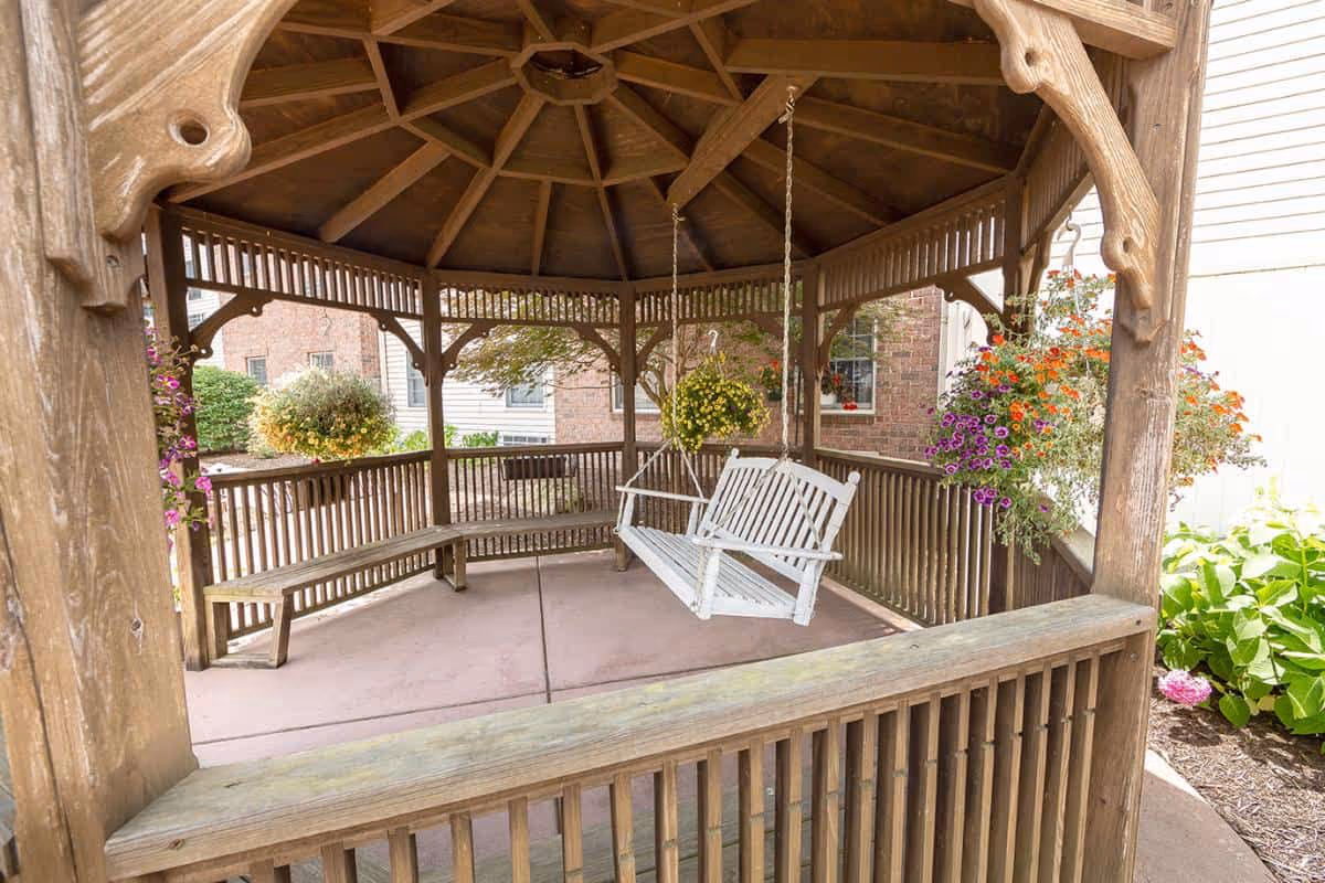Wooden gazebo with a white swinging bench inside, surrounded by hanging flower baskets and garden plants, located outdoors near a building.