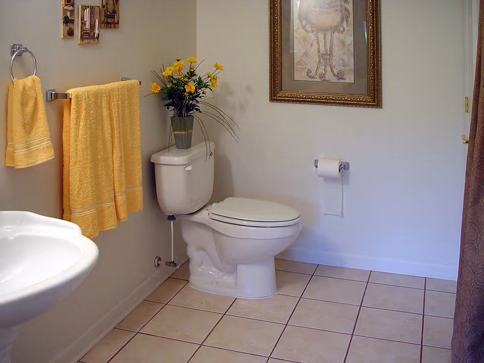 A clean bathroom with a white toilet and sink, yellow towels hanging on a towel rack, a vase with yellow flowers on top of the toilet tank, a framed picture on the wall, and a roll of toilet paper on a holder.
