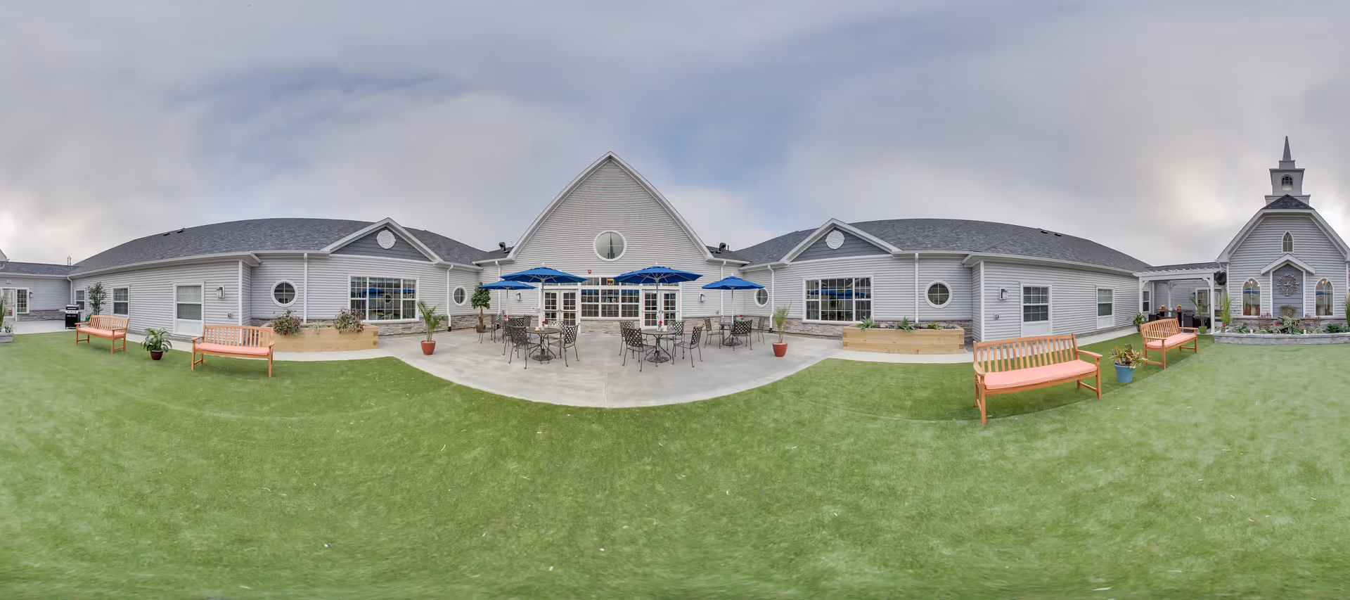 Outdoor courtyard area of Peregrine Senior Living at Colonie featuring a large green lawn, several wooden benches, potted plants, and a concrete patio with tables and chairs shaded by blue umbrellas. The building surrounding the courtyard has light gray siding, multiple windows, and a small chapel with a steeple on the right side under a cloudy sky.