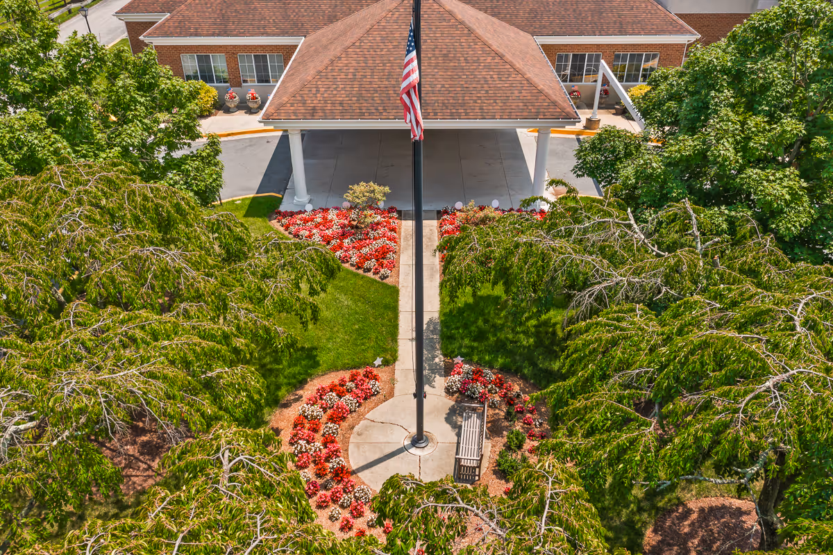 Aerial view of the entrance to Autumn Lake Healthcare at Silver Spring, showing a covered driveway with a peaked roof supported by white columns. In front of the entrance is a flagpole with an American flag, surrounded by a circular flower bed with red and white flowers and a bench. The area is landscaped with green grass and trees.