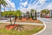 Exterior view of Veranda Club senior living facility with a white building, palm trees, landscaped garden beds with red mulch, and a clear blue sky with scattered clouds.