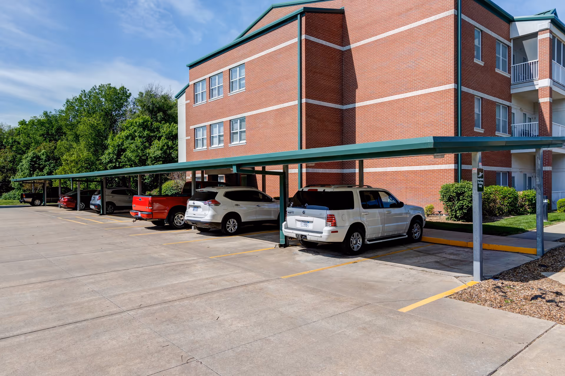 Parking area with a covered carport next to a three-story brick building with multiple windows. Several cars and trucks are parked under the green metal roof structure. Trees and greenery are visible in the background under a clear blue sky.