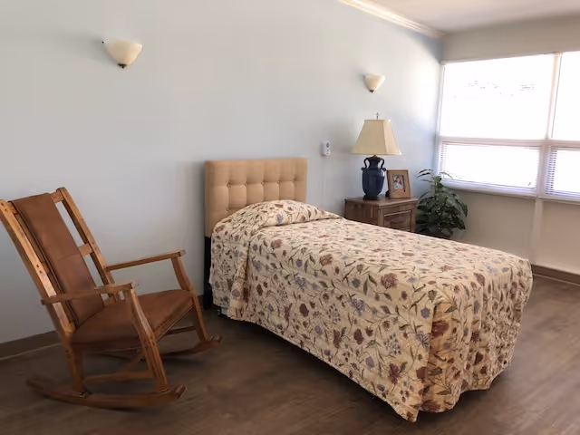A simple bedroom with a single bed covered with a floral patterned bedspread, a wooden rocking chair, a wooden nightstand with a blue lamp and a framed photo, and a large window letting in natural light.