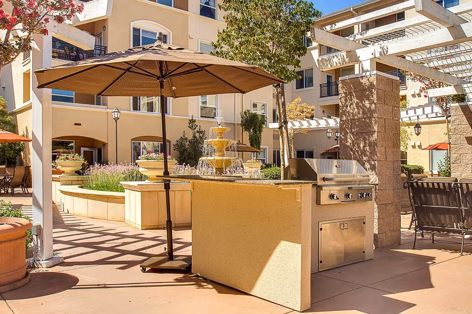 Outdoor patio area at Heritage Estates featuring a built-in stainless steel grill, large brown patio umbrella, seating area with chairs, decorative fountain, potted plants, and surrounding multi-story building with balconies.