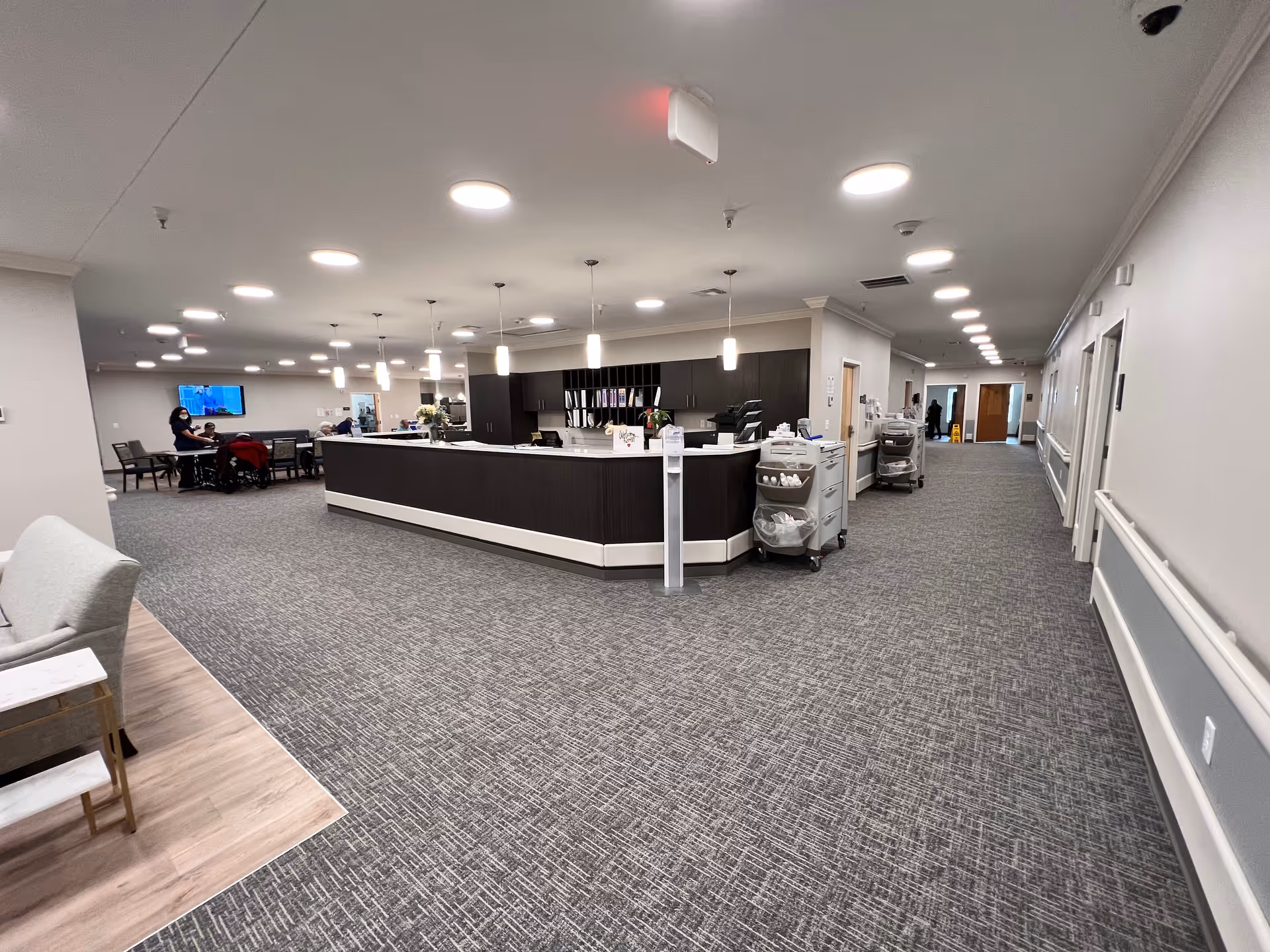 Interior view of a senior living facility reception area with a large dark wood reception desk in the center, overhead pendant and ceiling lights, gray patterned carpet, and a hallway leading to rooms on the right. In the background, a few people are seated near a television mounted on the wall.