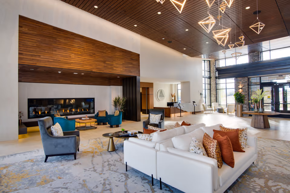 A spacious and modern living room area in The Ridge Pinehurst facility featuring a large white sectional sofa with multiple decorative pillows, several armchairs, a long horizontal fireplace set into a wooden accent wall, and large windows allowing natural light to fill the space. The ceiling has wooden paneling with contemporary geometric pendant lights hanging down. There is a round table with vases of flowers near the entrance.