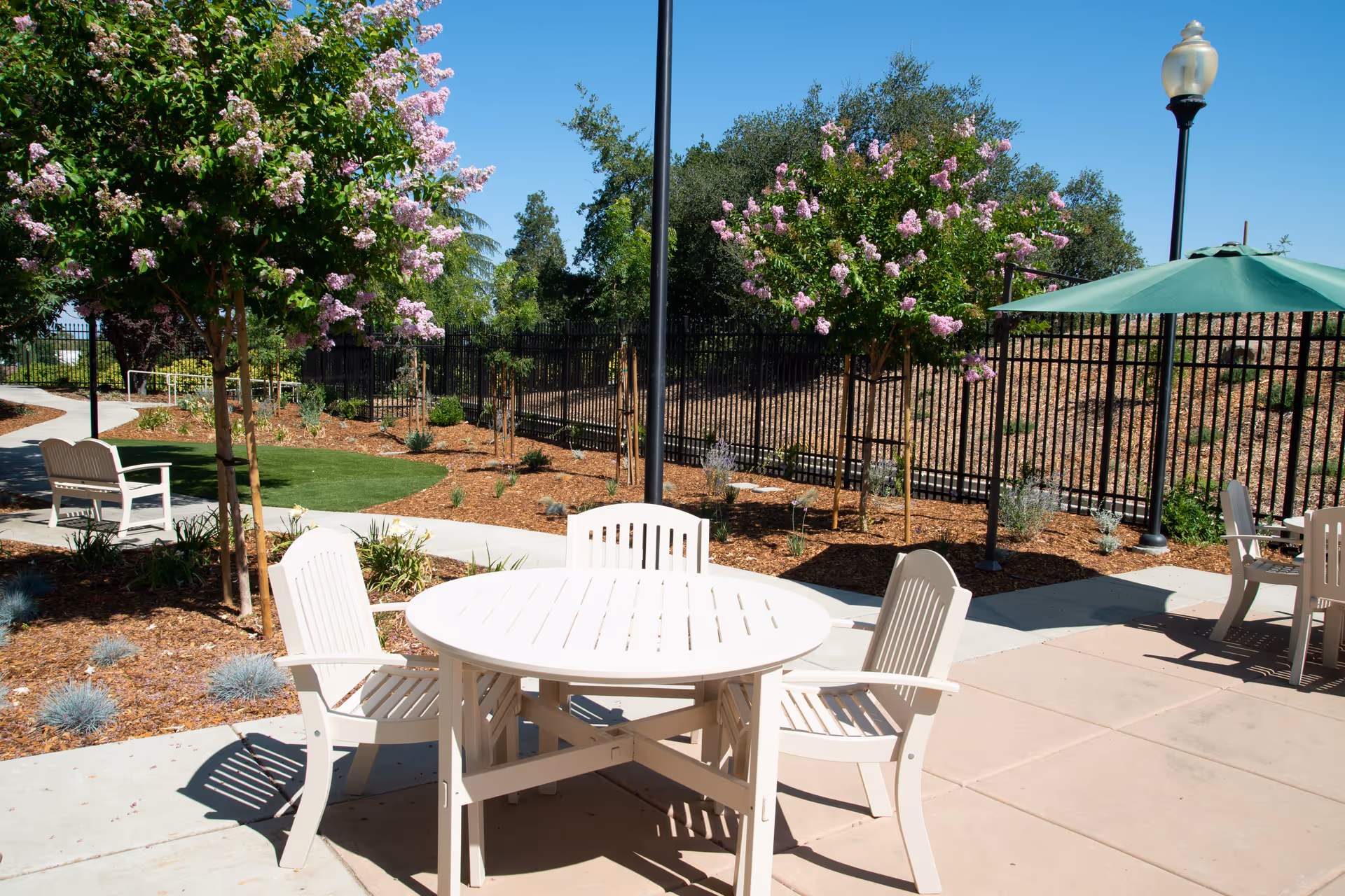 Outdoor patio area with white round table and four chairs, surrounded by flowering trees and landscaping under a clear blue sky. There is a green umbrella shading another table and chairs in the background, with a black metal fence and lamp posts visible.