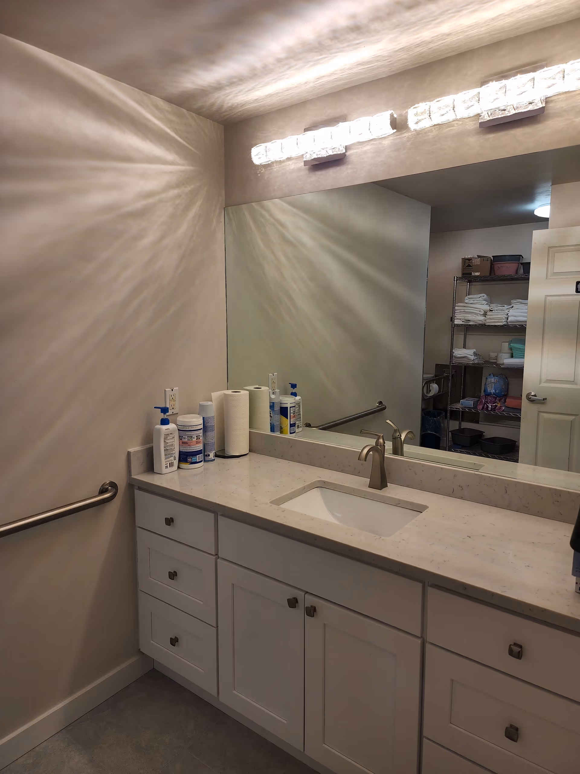 A bathroom vanity with a white countertop, a built-in sink, and a modern faucet. Above the sink is a large mirror reflecting a storage shelf stocked with towels and other supplies. The wall has a mounted light fixture with multiple bulbs casting decorative shadows. There is a grab bar on the left wall and various hygiene products on the countertop.