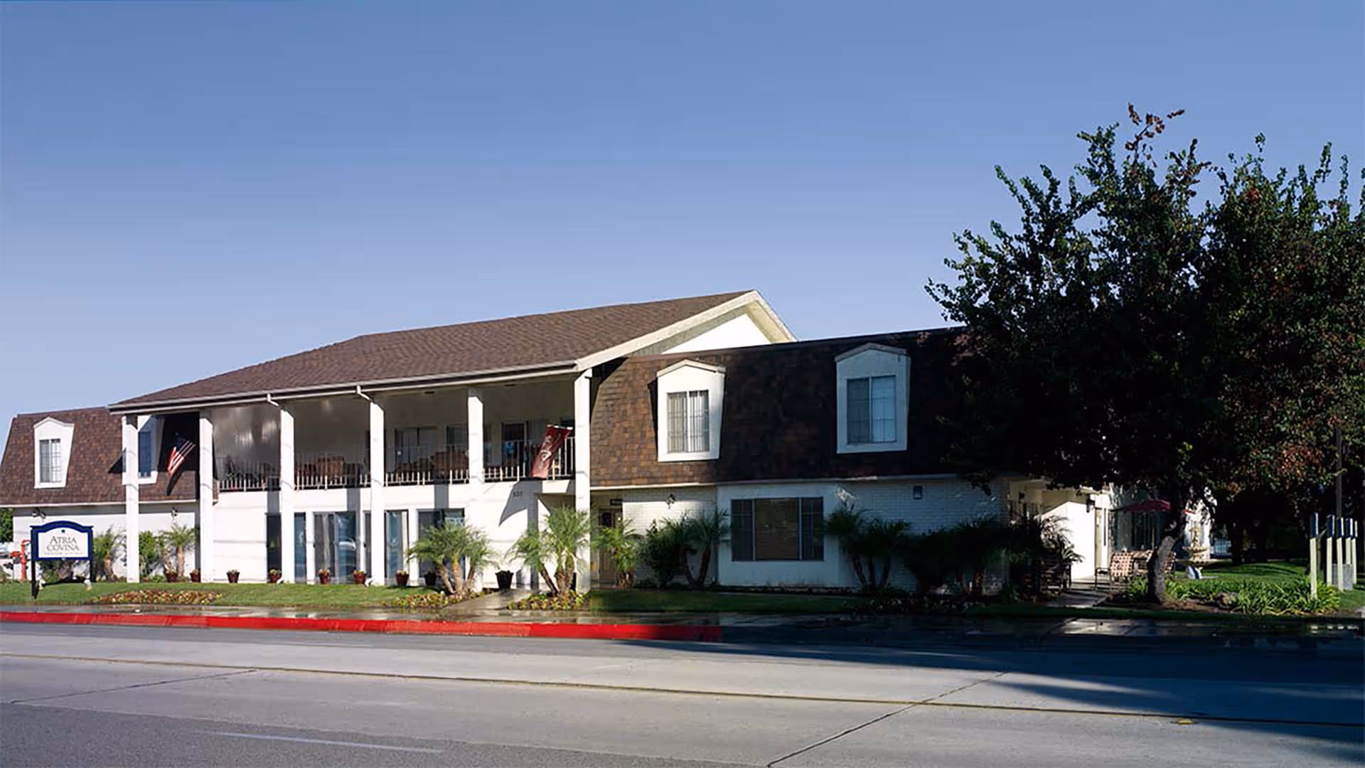 Exterior view of a two-story senior living facility building with a brown shingle roof, white walls, and a covered balcony on the second floor. There are several windows, palm plants, and a large tree on the right side. A sign near the street reads 'Atria Covina'.