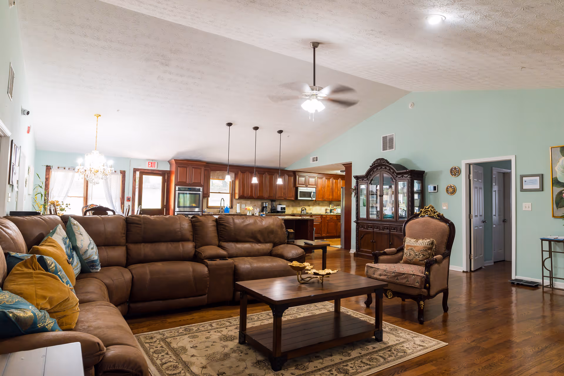Spacious living room with a large brown sectional sofa adorned with colorful pillows, a wooden coffee table on a patterned rug, an ornate armchair, and a dark wooden china cabinet. The room has light green walls, hardwood floors, ceiling fans, and an open view into a kitchen with wooden cabinets and pendant lighting.