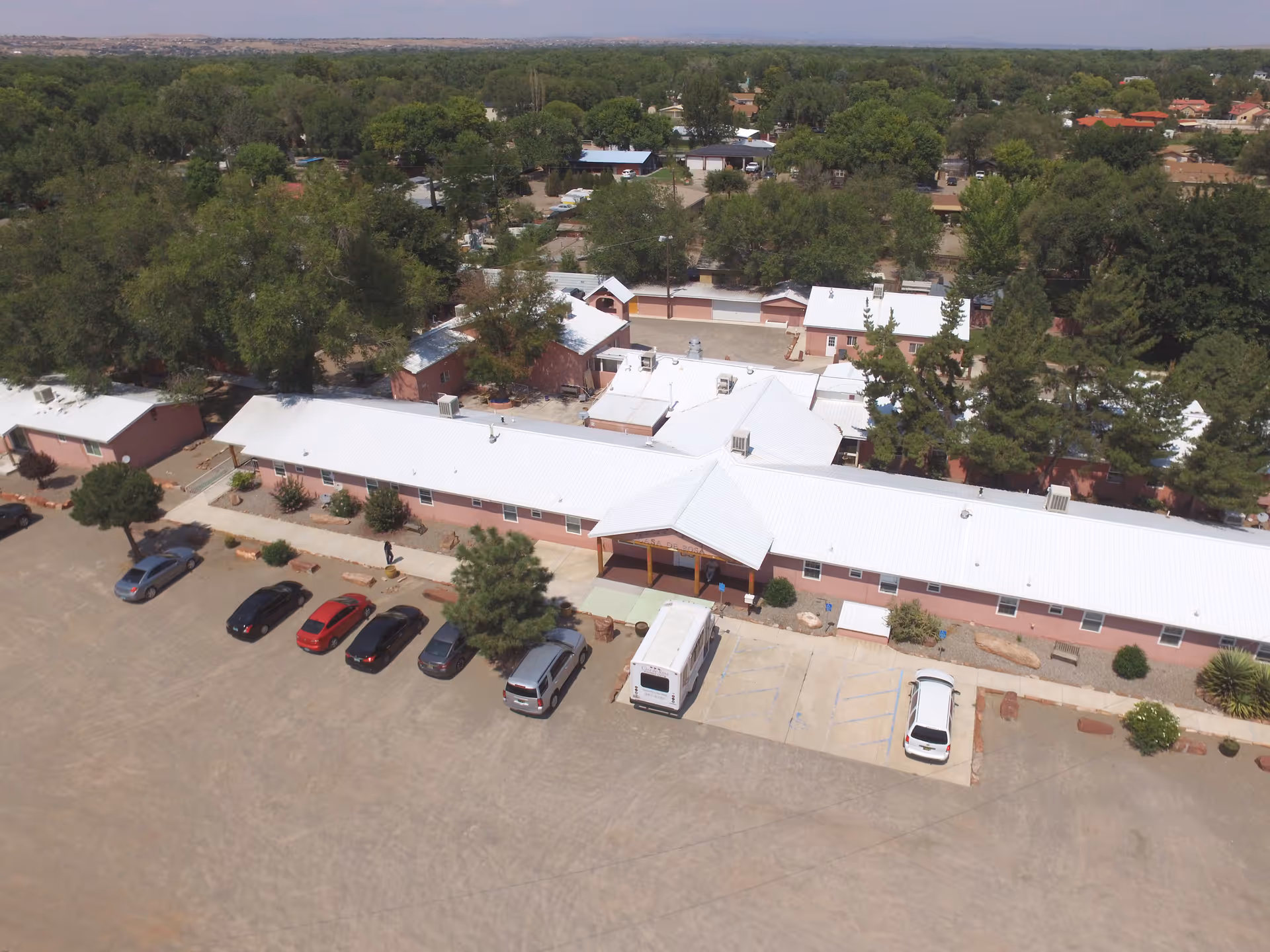 Aerial view of Casa de Rosa Assisted Living facility showing a long, single-story building with a white roof surrounded by trees and parked cars in front. The building is situated in a green area with many trees and other structures visible in the background.