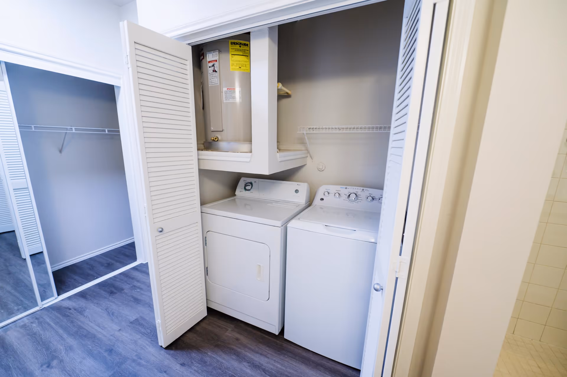 Laundry area with a white washing machine and dryer inside a closet with louvered doors. Above the machines is a water heater and a wire shelf. Adjacent to the laundry closet is a walk-in closet with mirrored sliding doors and a hanging rod.