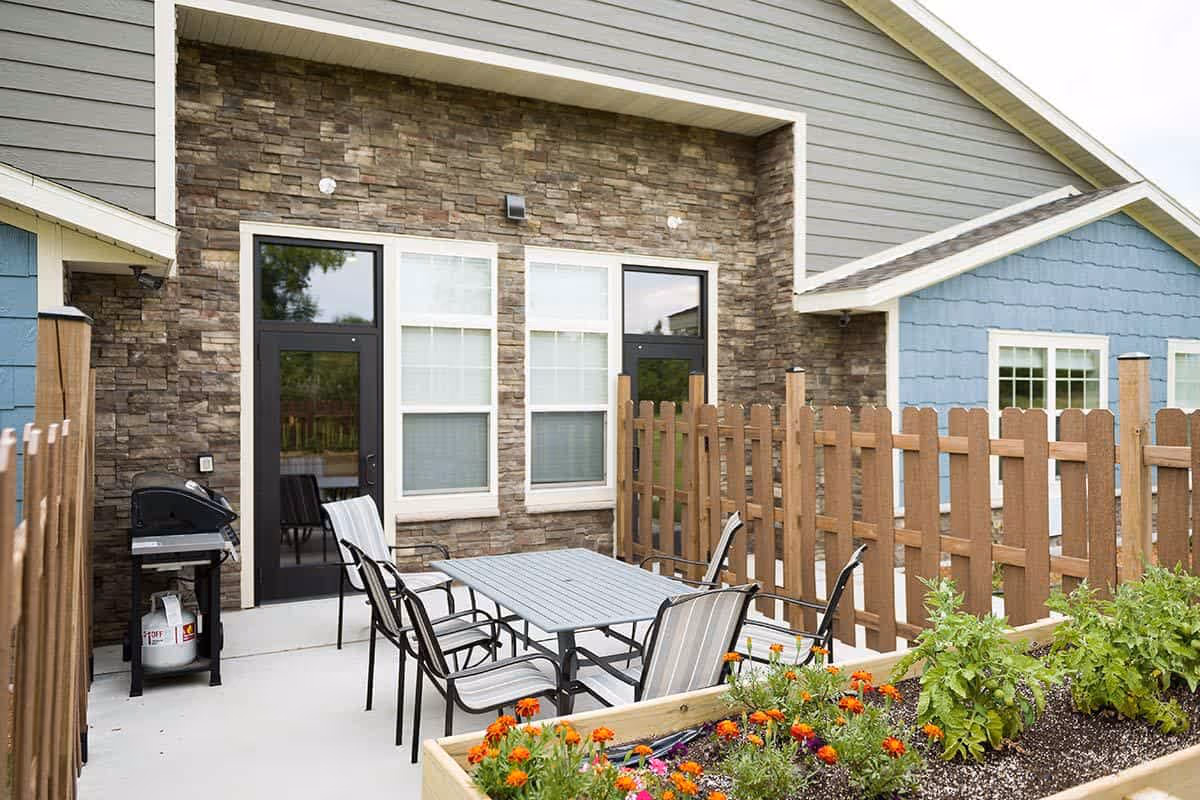 Fenced outdoor patio with a dining table and chairs, a grill, and raised flower beds beside a stone-clad building.
